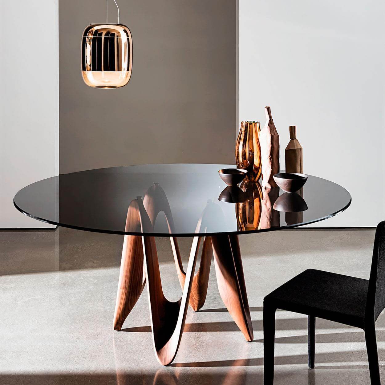 Round glass dining table with walnut sculptural base, Sovet Italia Cassoni copper light, vases, and black chair in modern setting.
