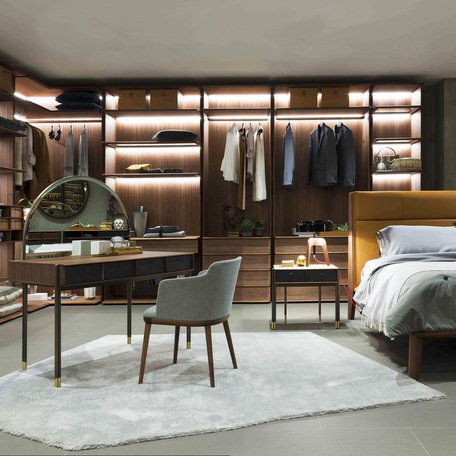 Walk-in closet with walnut veneer wardrobe, brass hardware, gray chair, mustard bed, and white linens in a modern transitional suite.