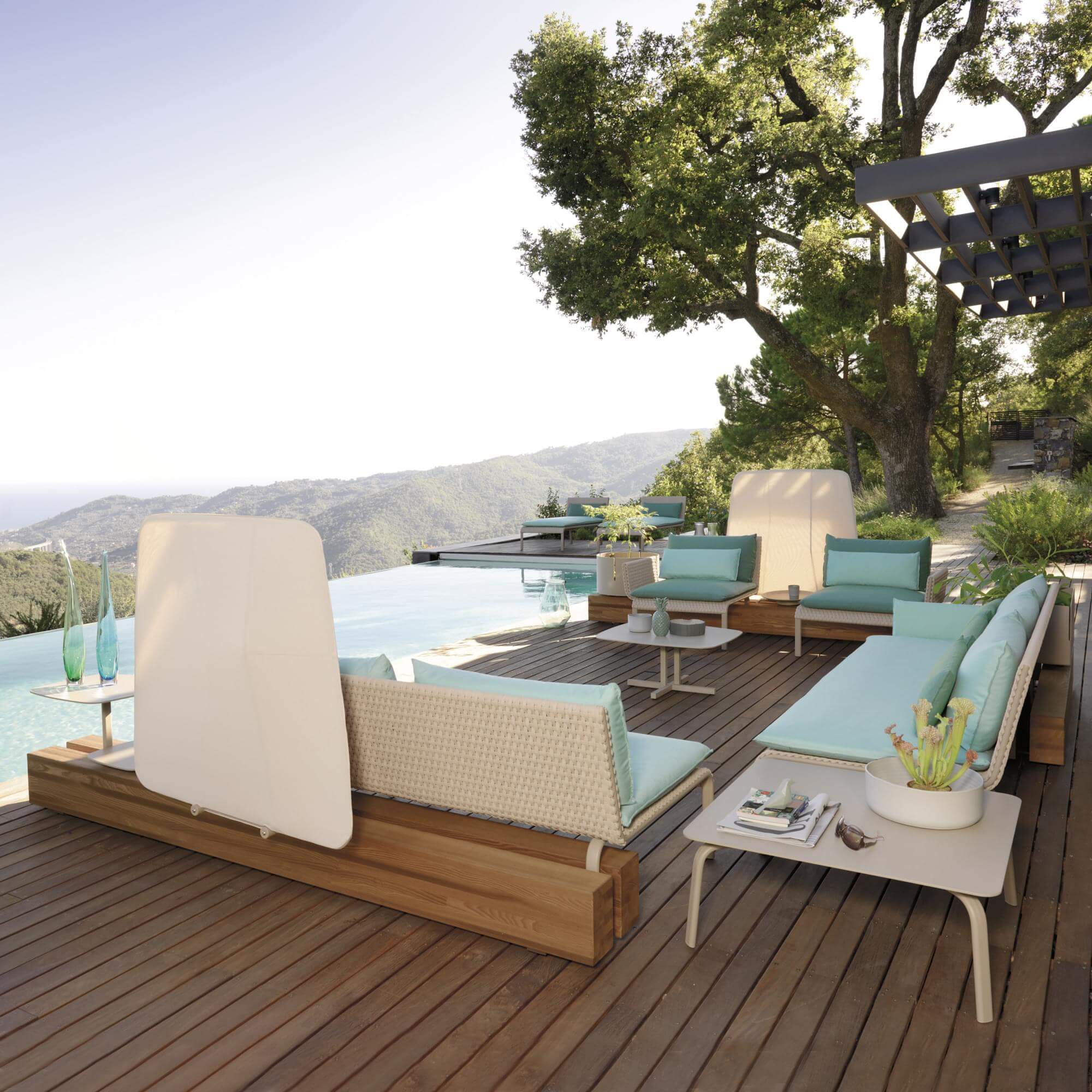 Outdoor lounge with woven rattan seating, turquoise cushions, pergola, wood deck, infinity pool, ocean view, glass sculptures.