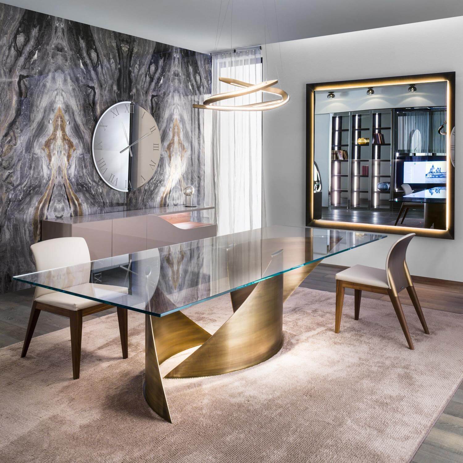 Glass dining table with flared brass base, beige Reflex chairs, marble wall, brass clock, and multi-tiered chandelier in a modern room setting.