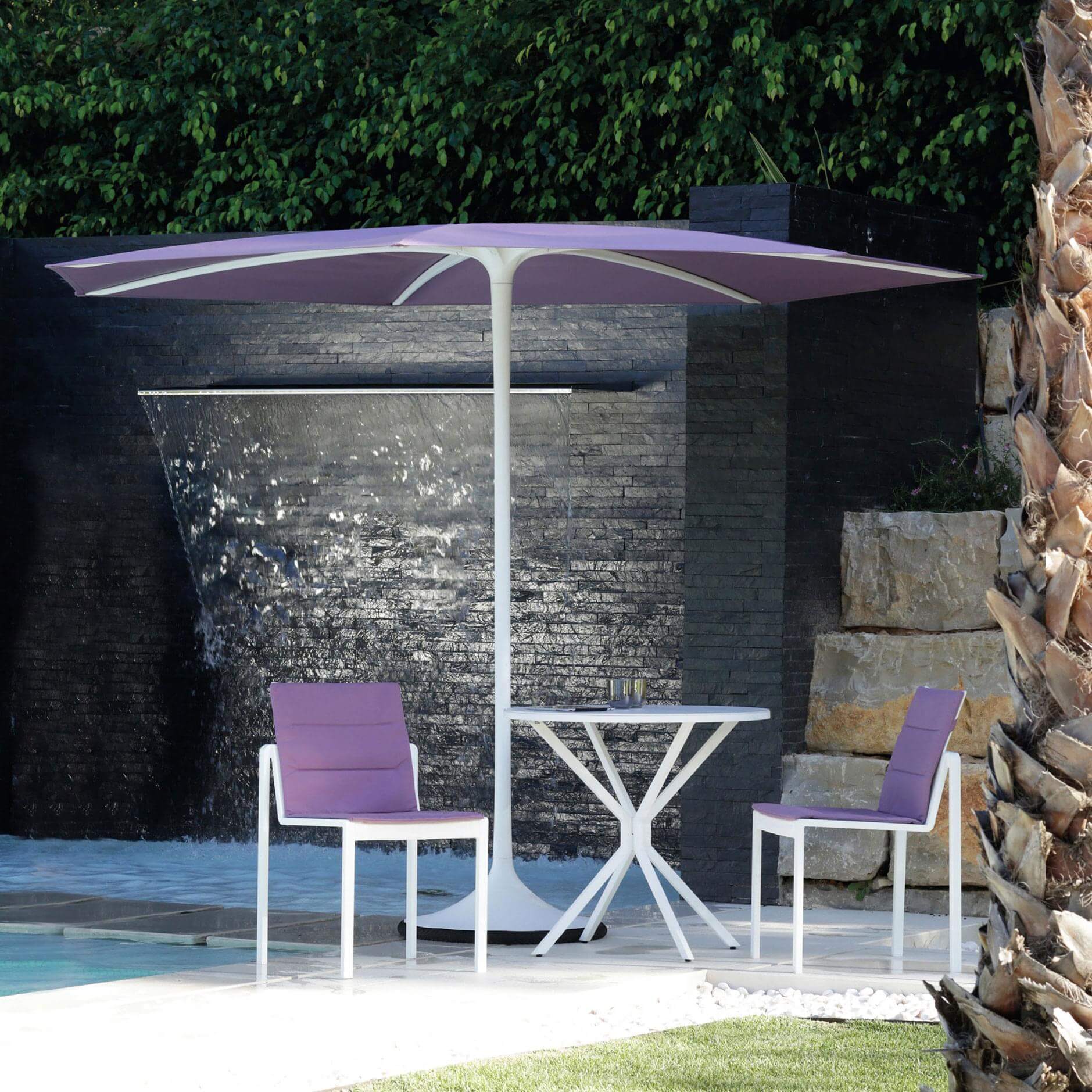 Lavender outdoor seating set with cantilever umbrella, round table, and mesh chairs, against a stone wall water feature.