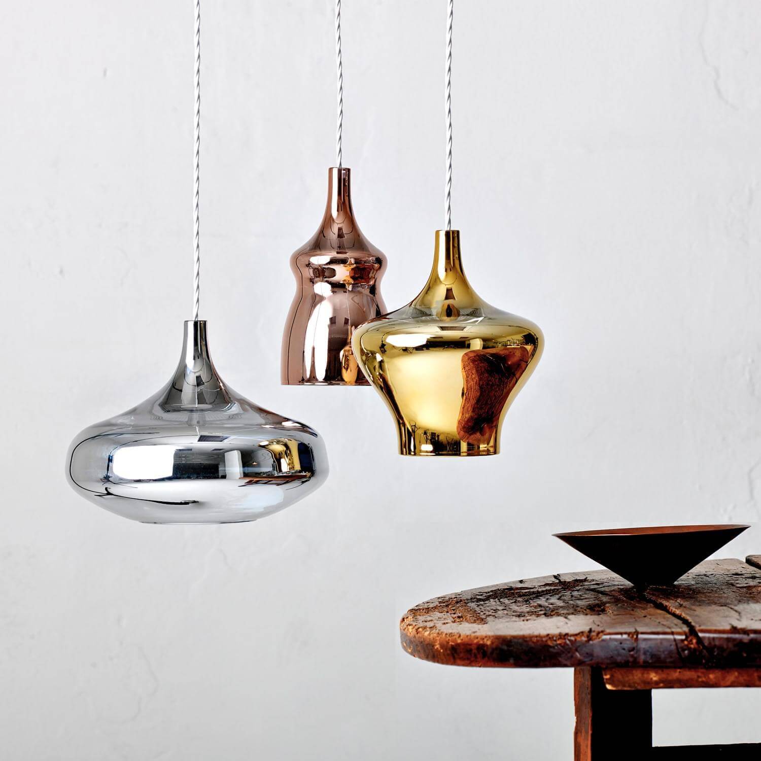 Pendant lights, silver, rose gold, gold finish, teardrop shape, white cords, against a white wall, with a dark wood table visible.