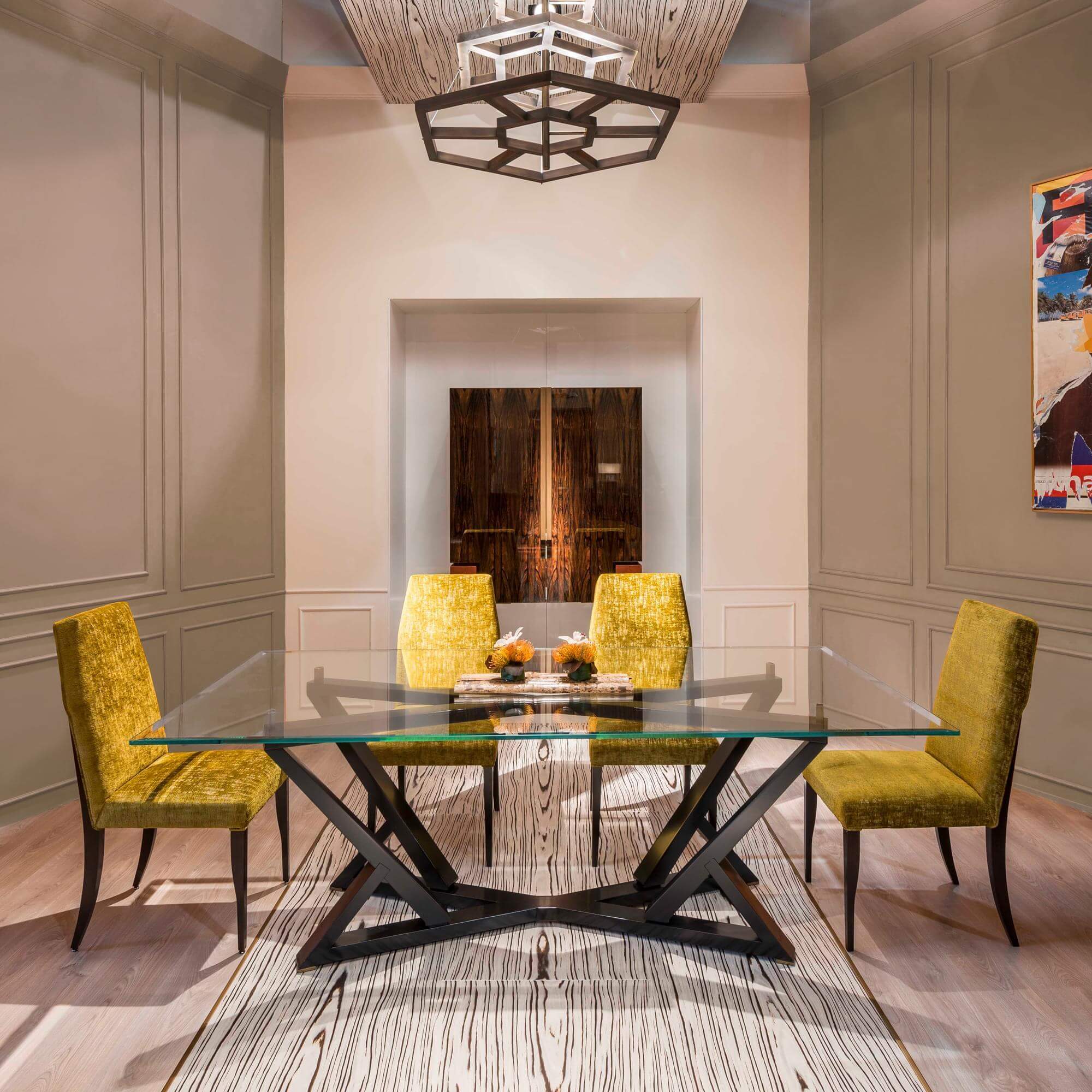 Dining room with glass table, mustard chairs, black metal base, beige walls, geometric chandelier, textured rug, and abstract artwork.