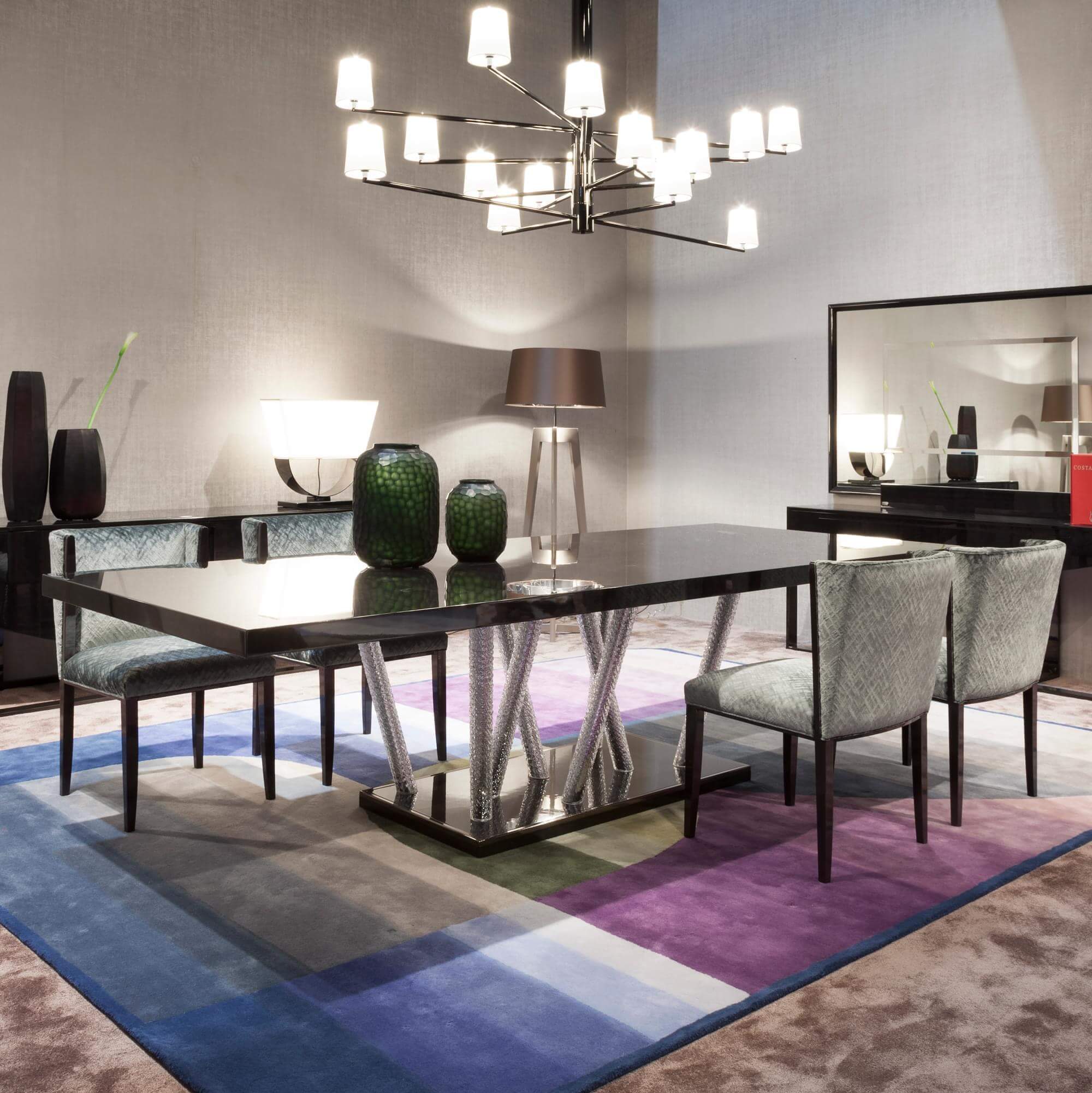 Rectangular dining table with black reflective surface and silver twisted metal base, centered on a multi-color patchwork area rug with gray chairs.