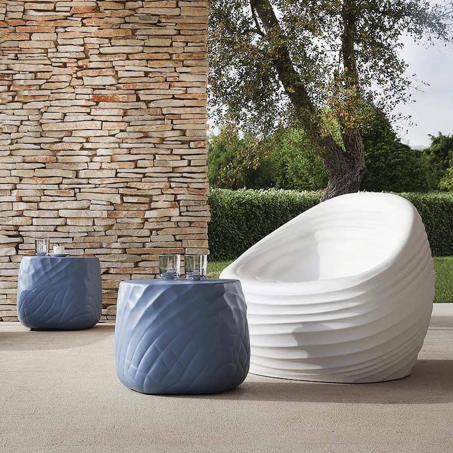White sculptural armchair with wave-like design, beside gray quilted side tables on beige patio with stone wall backdrop.