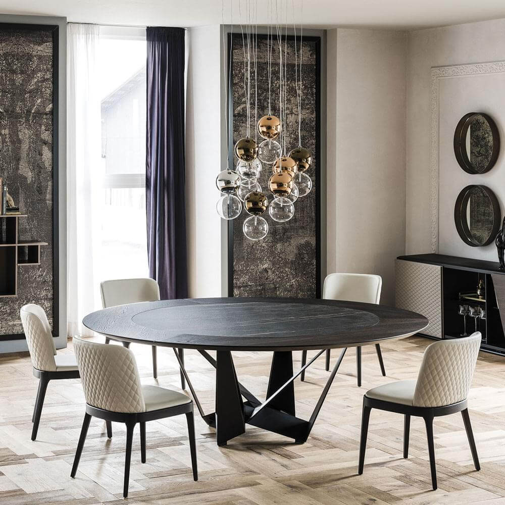 Dining table with dark gray top and black metal base, surrounded by four light beige quilted chairs, under a gold spherical chandelier.