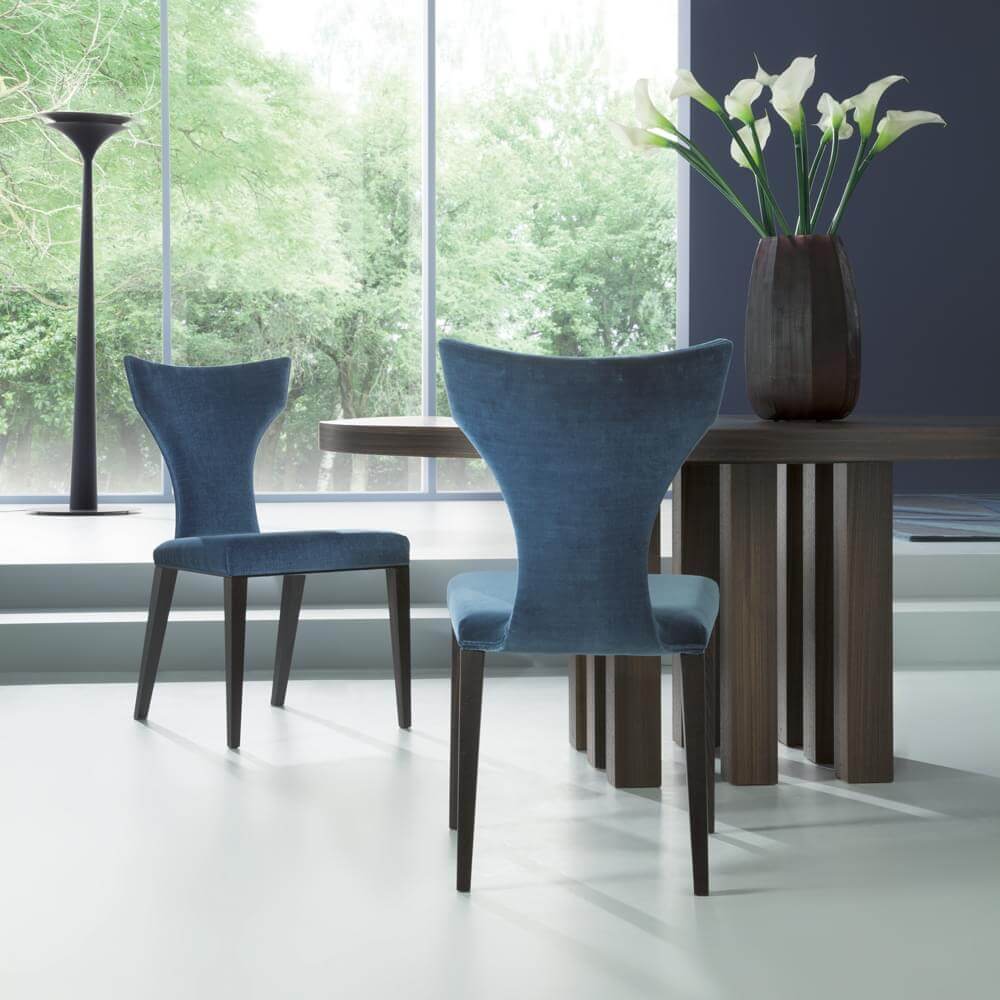 Teal velvet dining chairs with sculpted backs and flared wings sit with a dark wood rounded table in a modern blue interior.