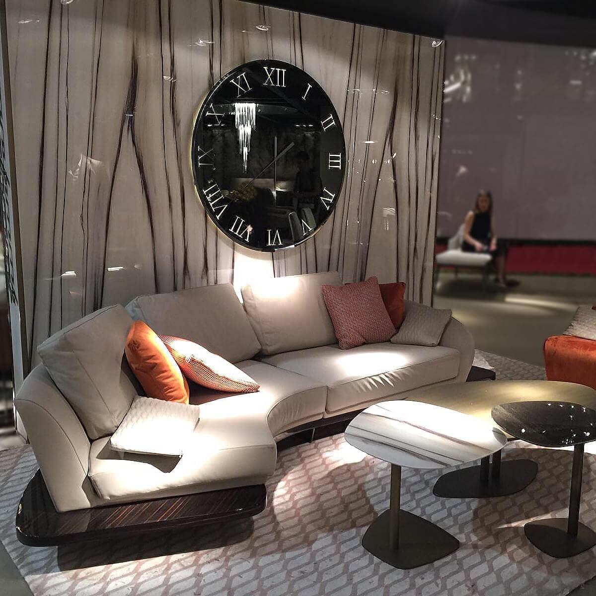 Curved off-white sofa with orange pillows, dark coffee tables on geometric rug, black wall clock, paneled wall, showroom setting.