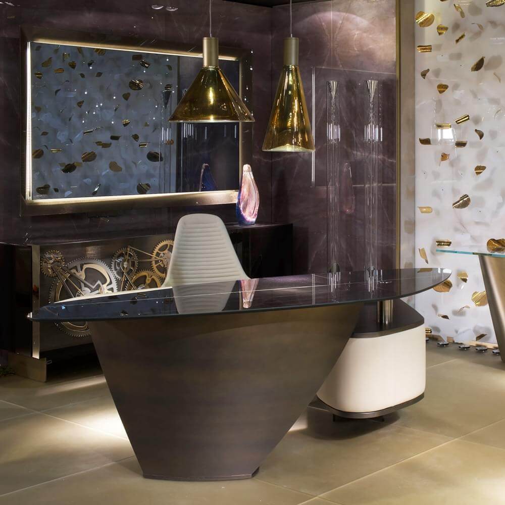 Curved desk with glass top, dark brown pedestal, gold pendant lights, gear display, gold-accented textured walls, purple backdrop, beige tile floor.