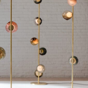 Floor lamps with brass stems and multi-colored glass globes arranged against a white brick wall in a cascading display.