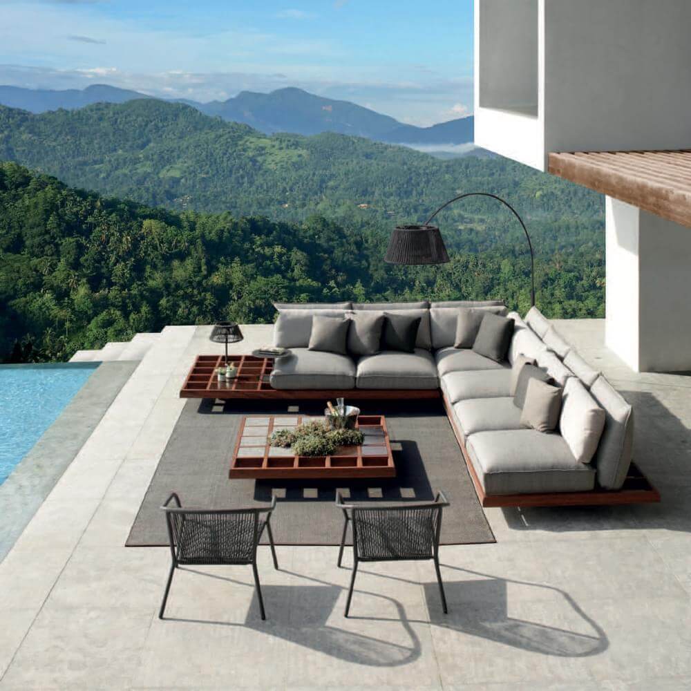Outdoor lounge with light gray sectional sofa, dark wood frame, black chairs, moss-green coffee table, tiled patio, and hillside view.