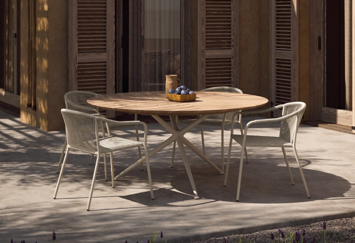 Round dining table with light wood top and white metal star base, surrounded by four white rope seat chairs on concrete patio.