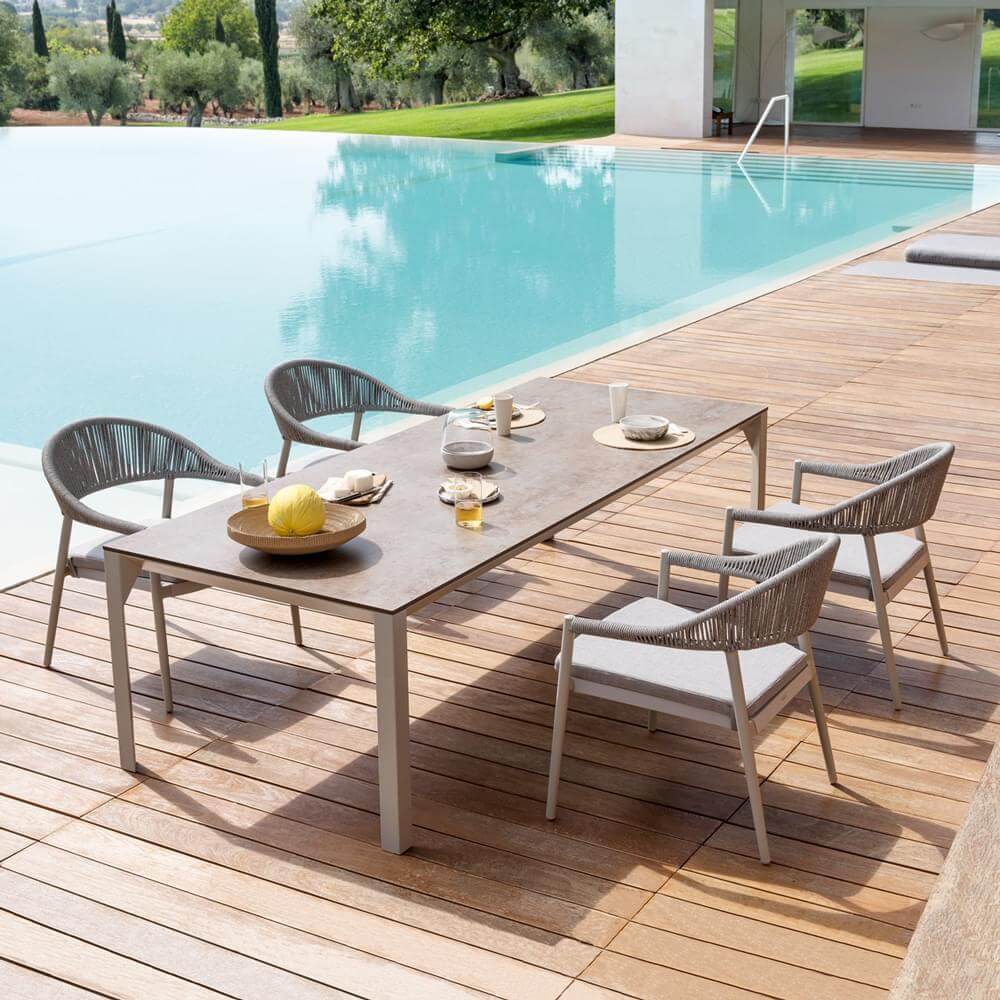 Dining set with dark brown textured table, light gray metal legs, woven gray rope armchairs, and turquoise pool backdrop.