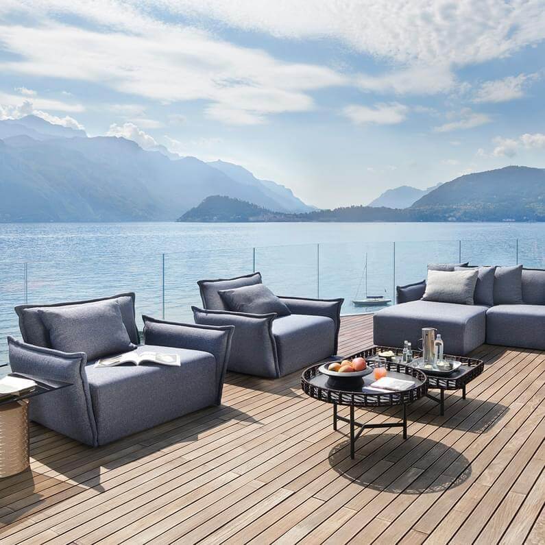 Outdoor sectional sofa and armchairs in gray textured fabric on a wood deck with a glass coffee table, overlooking a lake and mountains.
