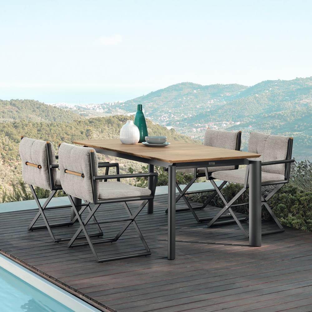 Outdoor dining table with teak top and gray metal frame, paired with gray fabric folding chairs featuring tan leather straps, on a wooden deck.