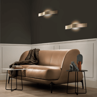 Tan leather sofa with Masiero Libe wall lamps, dark wood paneling, light wood coffee table, and stacked gray/beige decor.