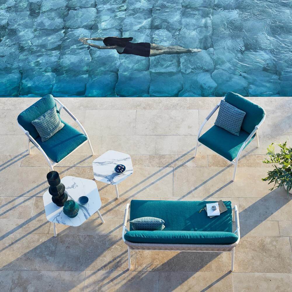 Outdoor seating group with teal rattan furniture, marble side tables, and beige stone tile flooring beside a turquoise swimming pool.
