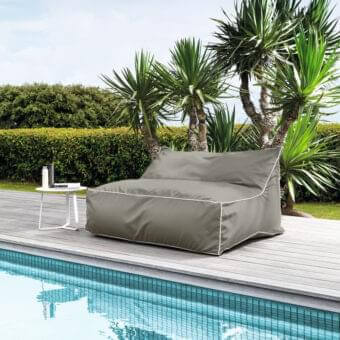 Gray cushioned rectangle sits beside a light blue tiled pool with plants and a white surface nearby