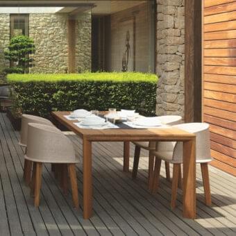 Rectangular table with six light chairs sits on gray planks before a stone wall and green foliage backdrop
