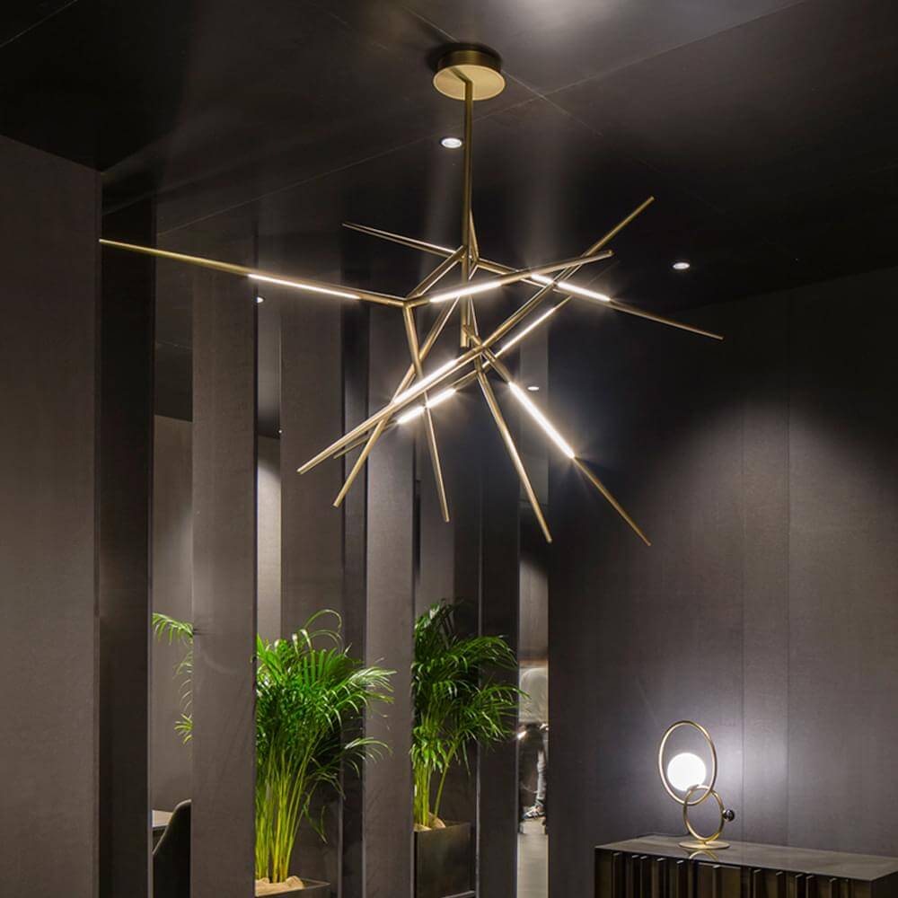 Modern chandelier with brass finish, radiating rods and integrated LED lights, suspended in a minimalist gray lobby with plants.