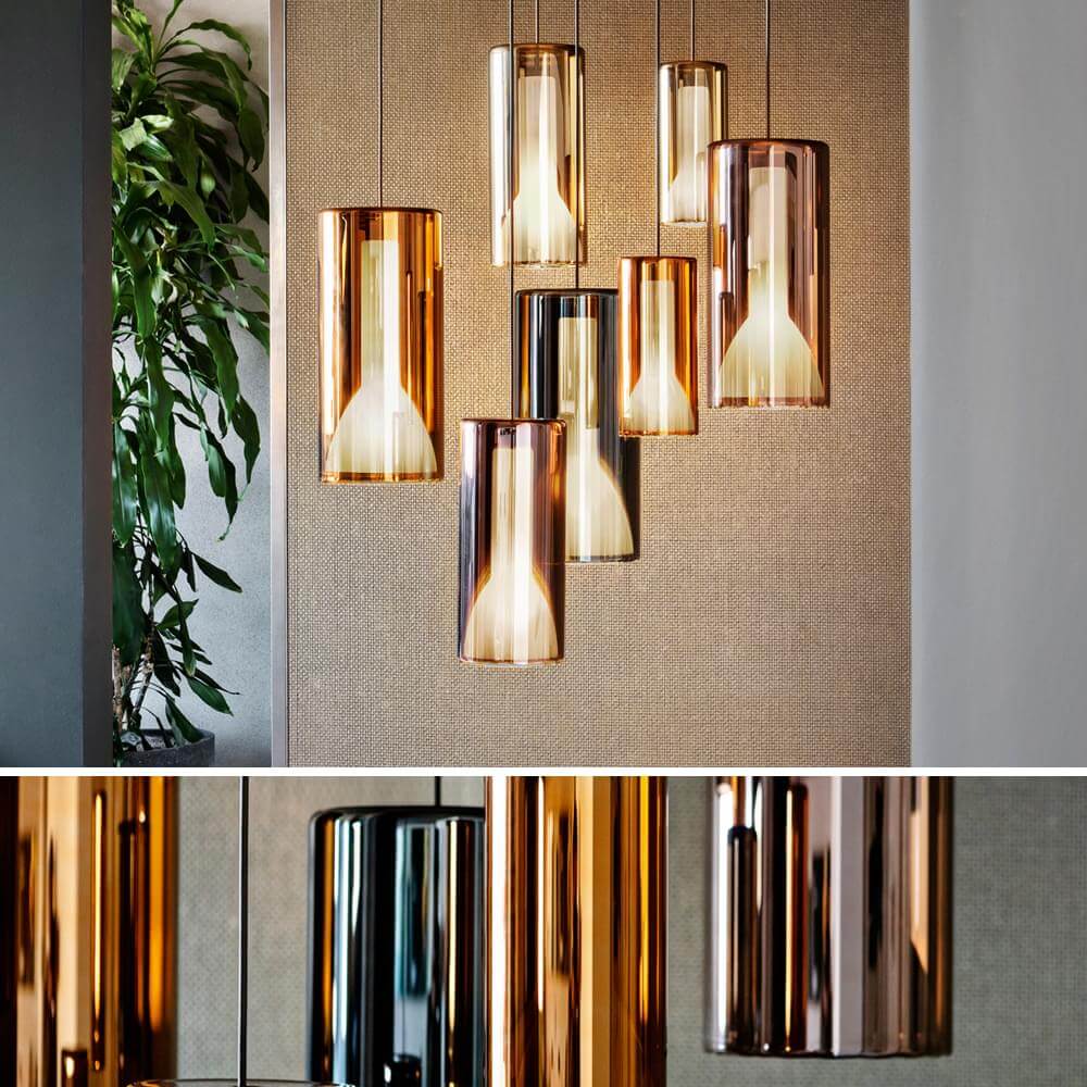 Pendant lights, amber glass, cylindrical shades with gradient color, suspended from dark cables, against a textured beige wall with plant.