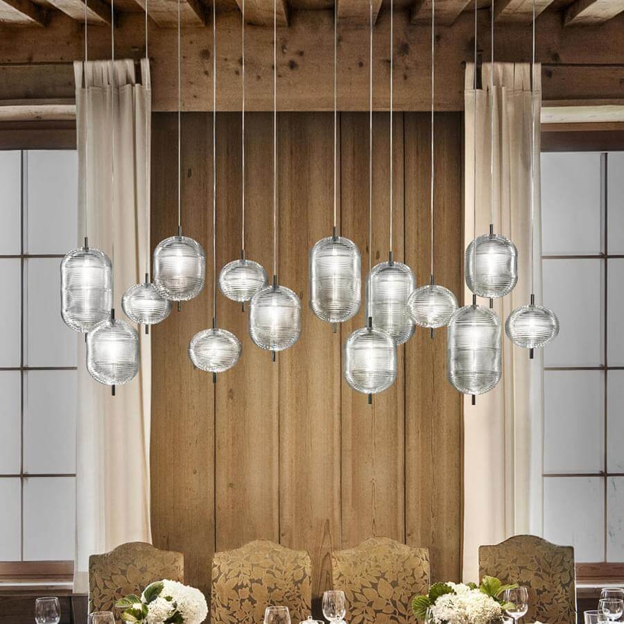 Fifteen-pendant light fixture with clear ridged glass shades and brushed metal rods hangs above a dining table in a rustic room with wood paneling.