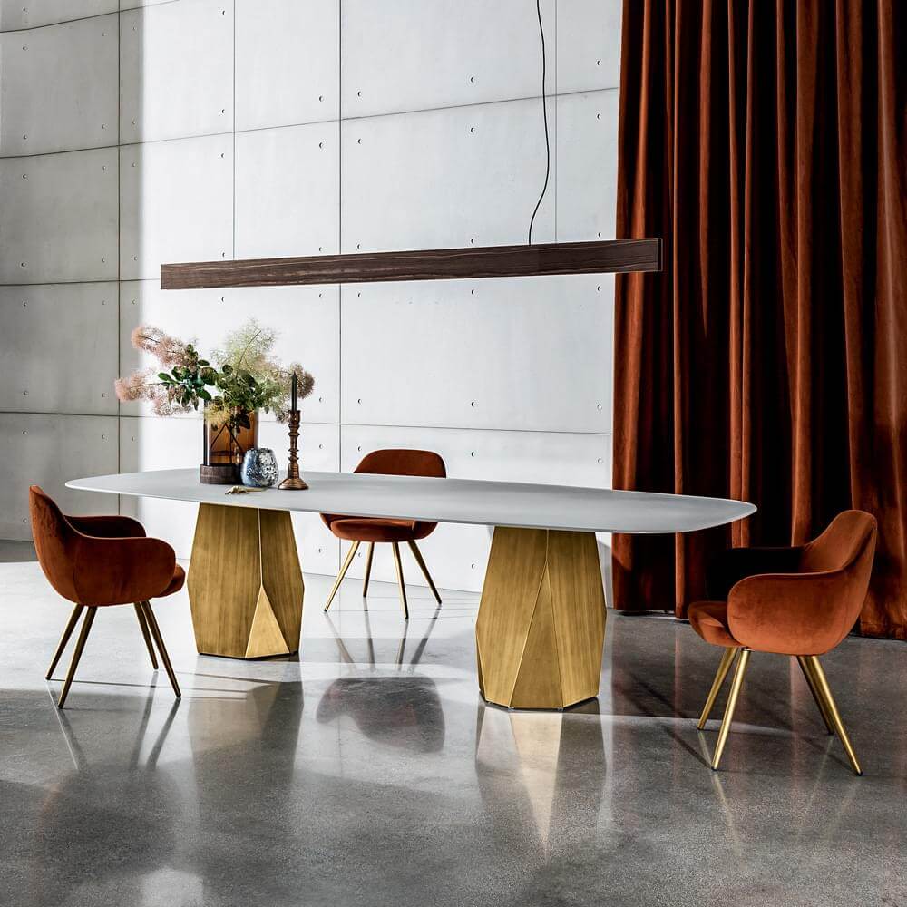 Dining table with white marble top and gold metal base, surrounded by terracotta velvet chairs, under linear lighting and rust curtain.