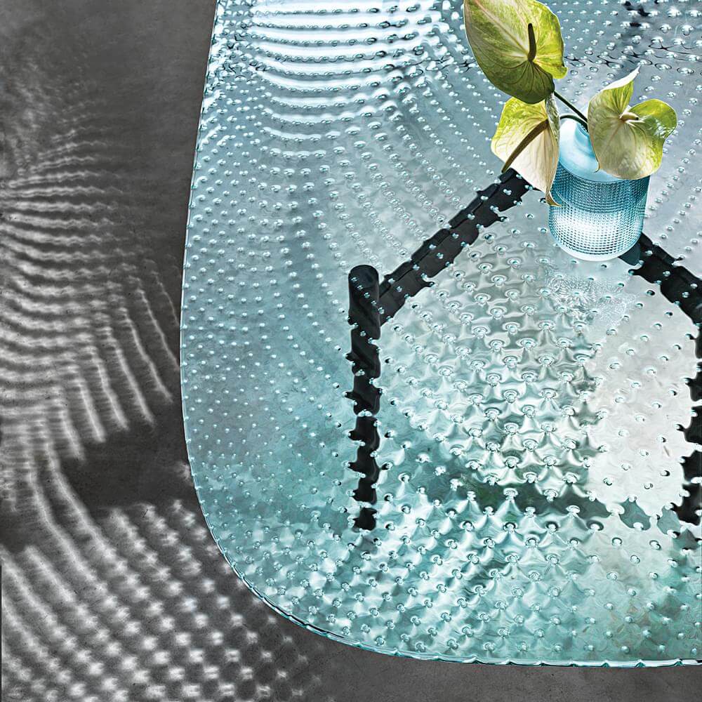 Aqua glass table with bubbled texture, black metal base, and orchid arrangement, against a gray wall.