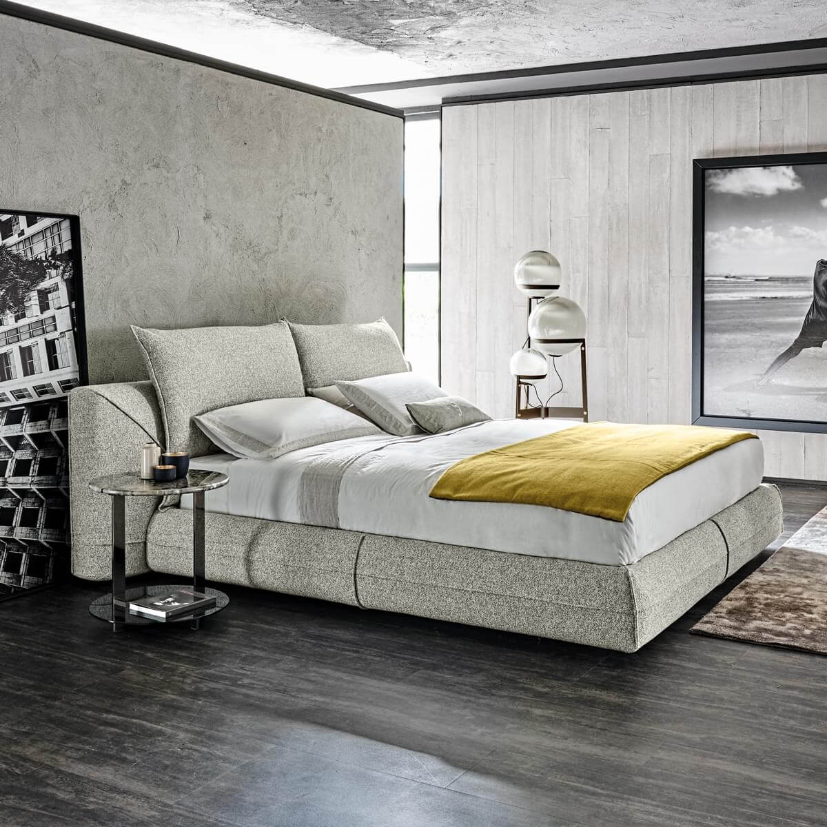Upholstered bed, light gray fabric, rectangular headboard with gray and white pillows, dark marble side table, textured plaster wall.