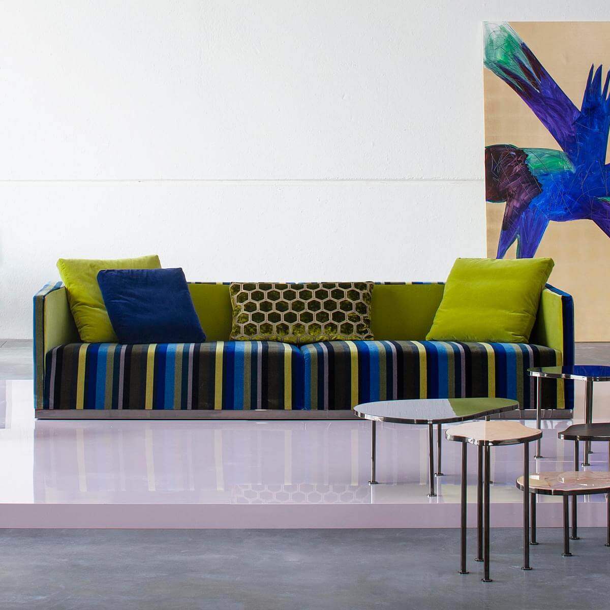Contemporary sofa with blue, yellow, black striped base, lime green back, velvet pillows, metallic side tables on pink floor.