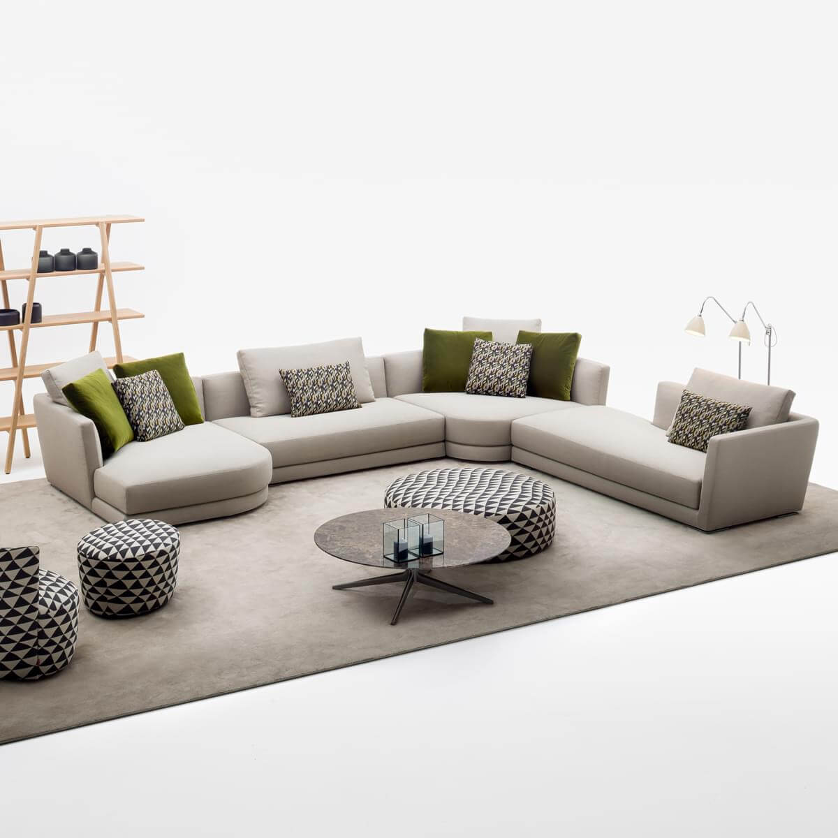Beige modular sectional sofa with L-shape chaise, olive & geometric pillows, glass coffee table, patterned ottoman & poufs, wood bookcase.