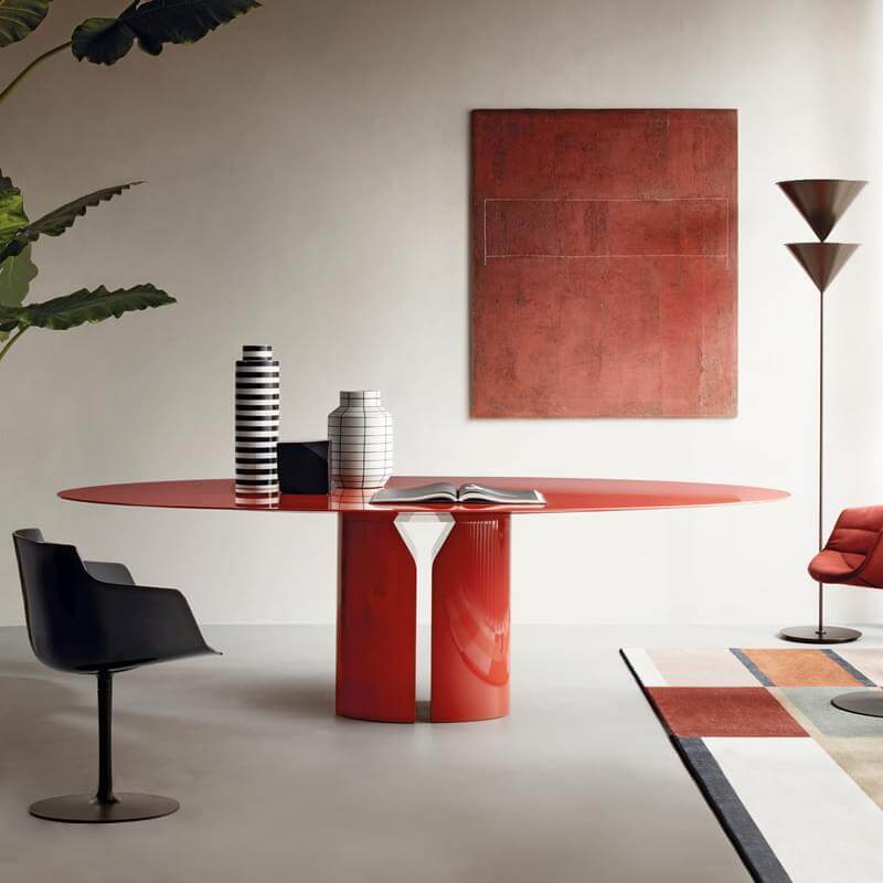 Red dining table with fluted pedestal, black plastic chair, leather chair, rust artwork, brass lamp, patterned rug, vases, book.