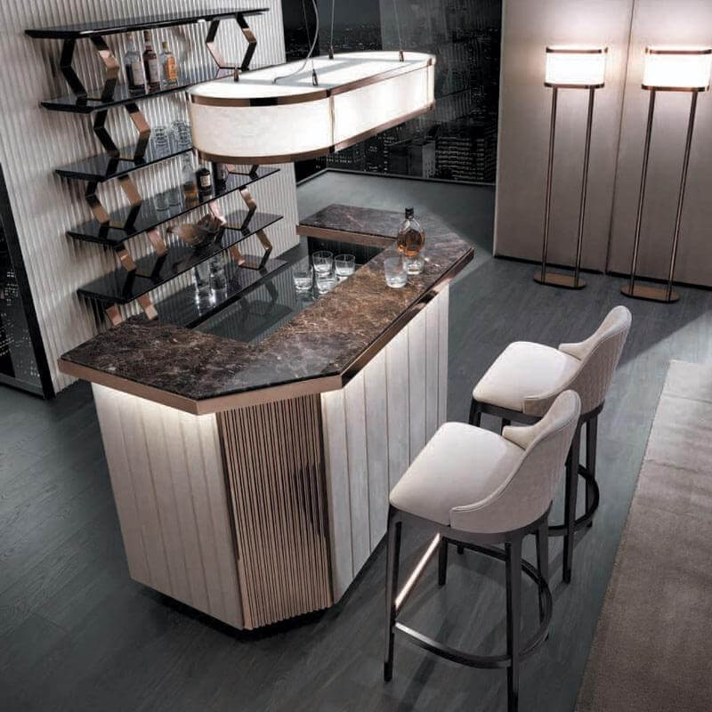 Bar with dark brown marble countertop, cream slatted base, and two beige upholstered stools, lit by brass fixtures.