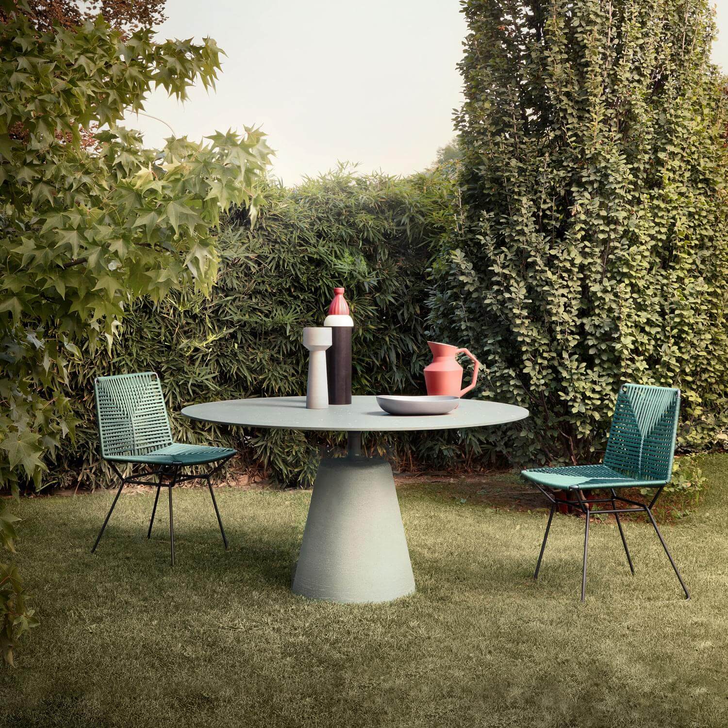 Sage green round dining table with turquoise cord chairs on lawn, featuring vase, pitcher, bowl, minimalist design, MDF Italia 2021.