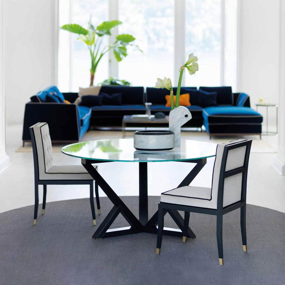Glass dining table with black metal X-base, gray & black armchairs, dark blue velvet sectional, patterned pillows, plant, and ceramic sculpture.