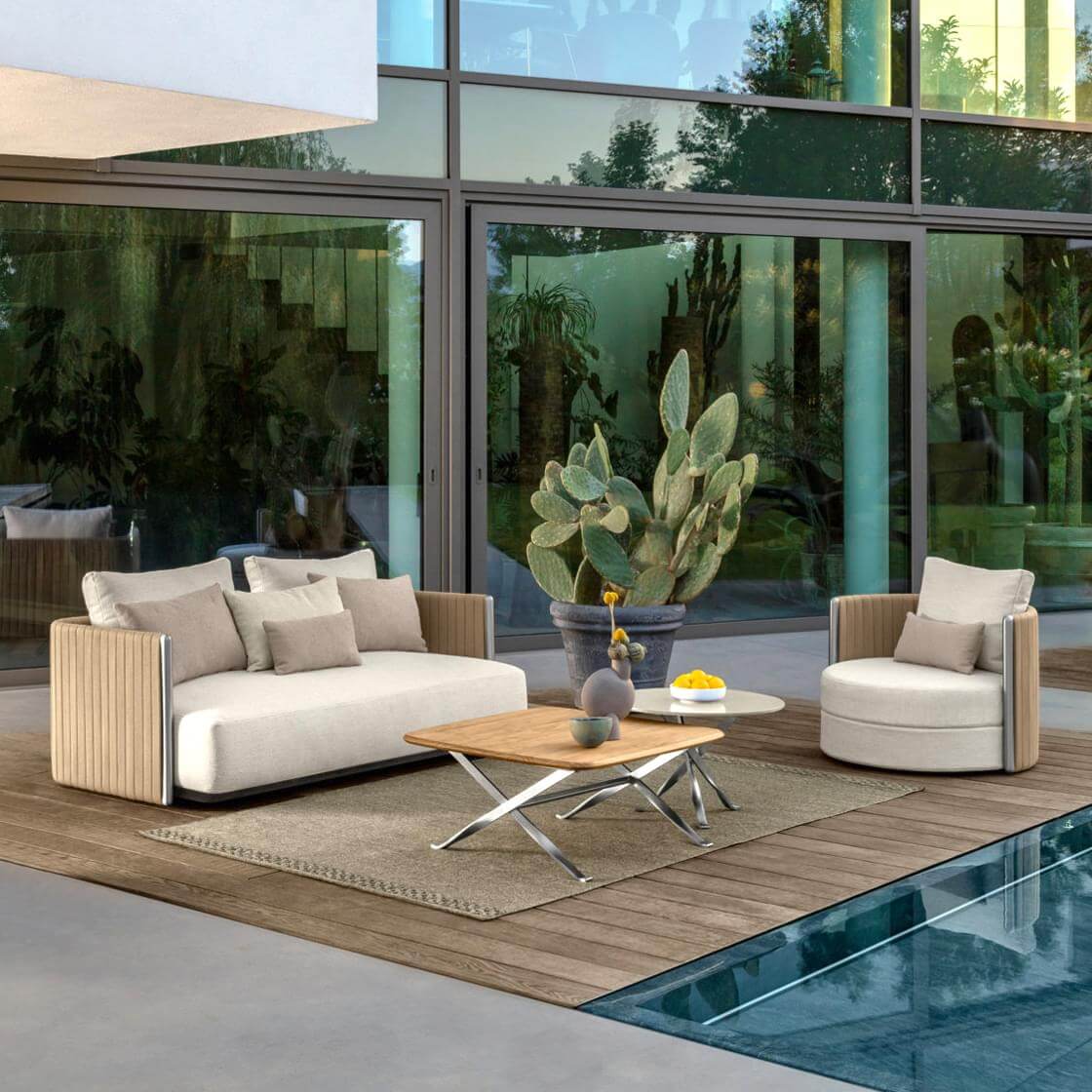 Beige outdoor sofa and armchair with woven sides, weathered wood coffee table with chrome base, on jute rug by pool.