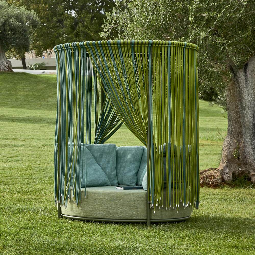 Outdoor seating with green rattan frame, circular shape, and fringed cord canopy, set on a green lawn under trees.