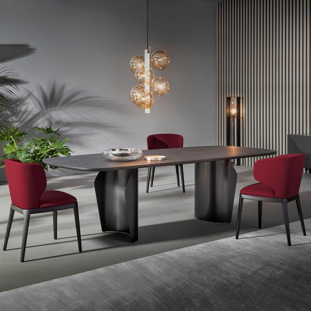 Dining table with dark gray textured top and curved base, paired with four red chairs, amber glass chandelier, gray rug, and wood paneling.