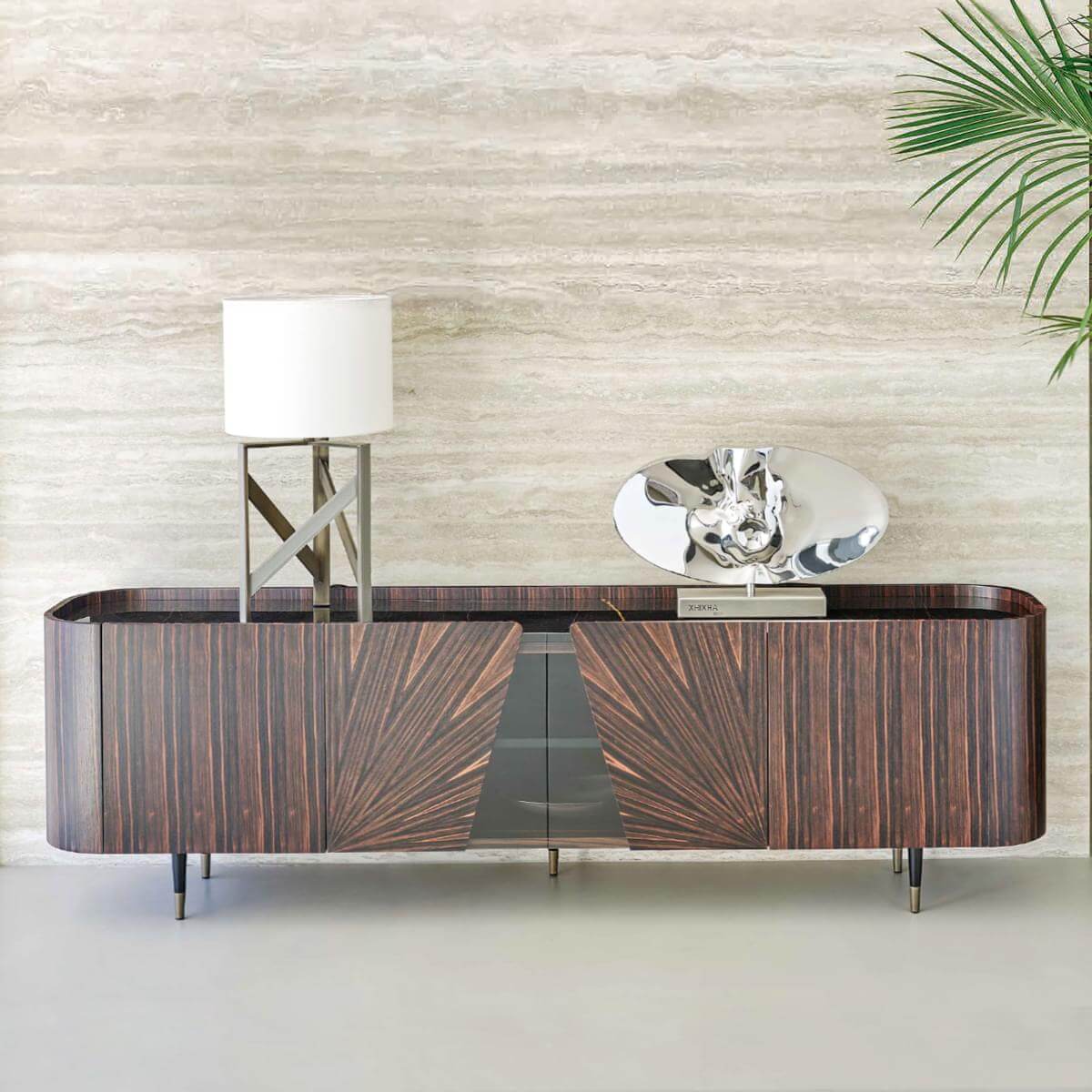 Dark wood console table with palm frond veneer, mirrored center, and metal legs, styled with lamp, mirror, book, and potted palm.