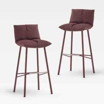 Two angled chair forms with curved backs and textured seats rest on slender metal frames against a pale backdrop