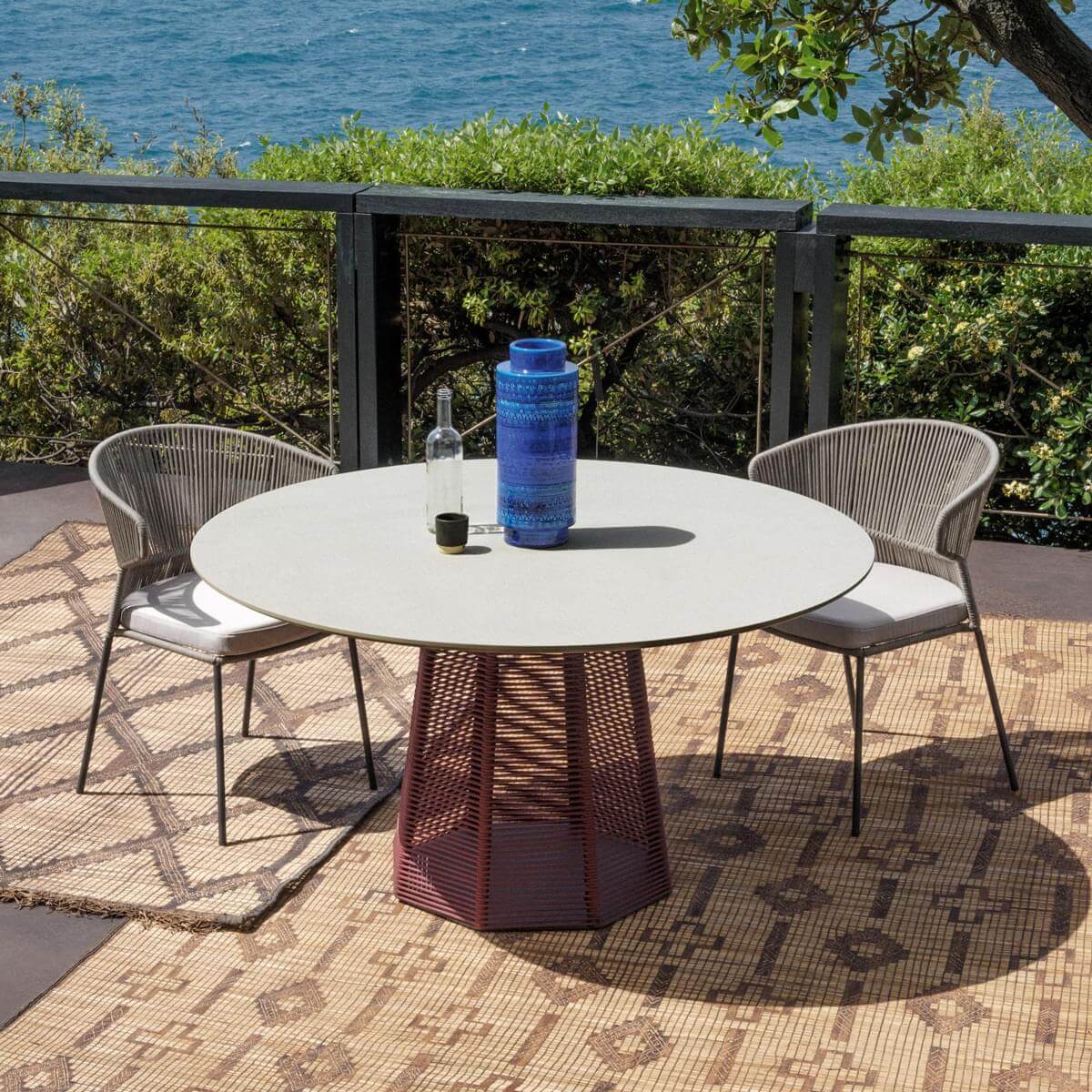 Round dining table with light gray matte top and burgundy woven pedestal, paired with gray woven chairs on a geometric beige rug.
