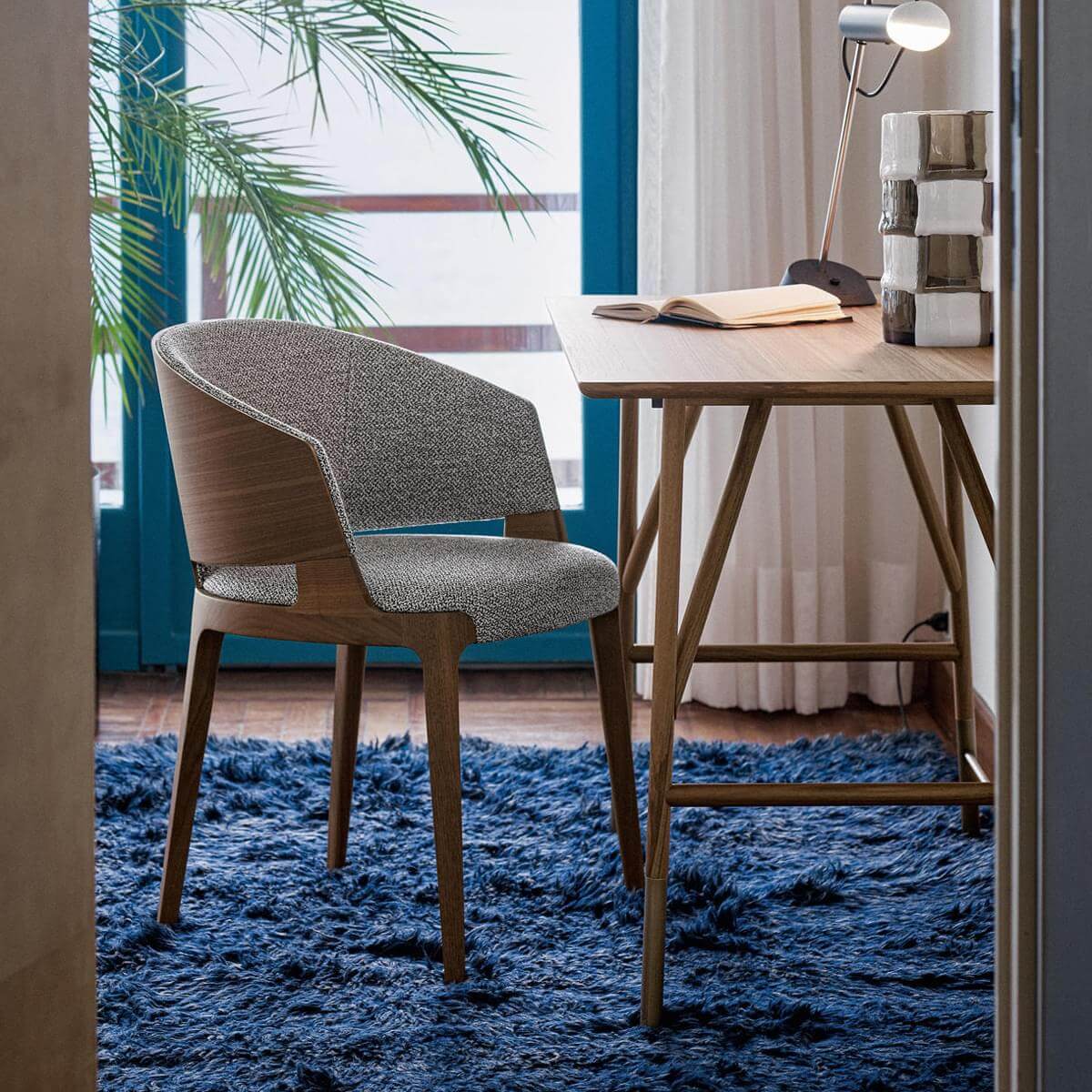 Dining chair with light wood frame and gray-beige textured seat on blue shag rug in modern room with desk, lamp, and palm.