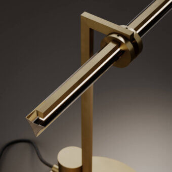 Brass table lamp with rectangular diffuser, cylindrical housing, geometric brass framework, and circular base, on a gray gradient backdrop.
