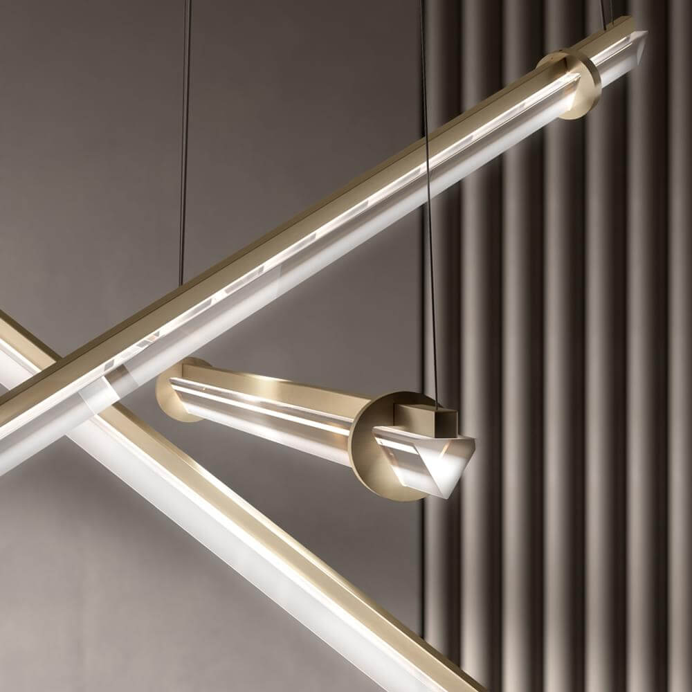 Modern chandelier with brushed gold metal frame, clear acrylic tubes, and faceted crystalline center, suspended against ribbed gray panels.