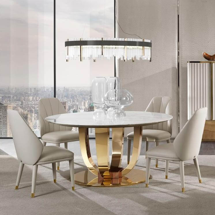 Round dining table with white marble top, gold metal base, and four beige upholstered chairs, under a cylindrical gold chandelier.