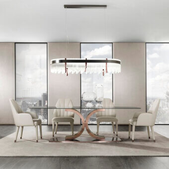 Rectangular glass dining table with bronze X-base, six beige fabric chairs, and modern chandelier over neutral rug in a bright dining room.