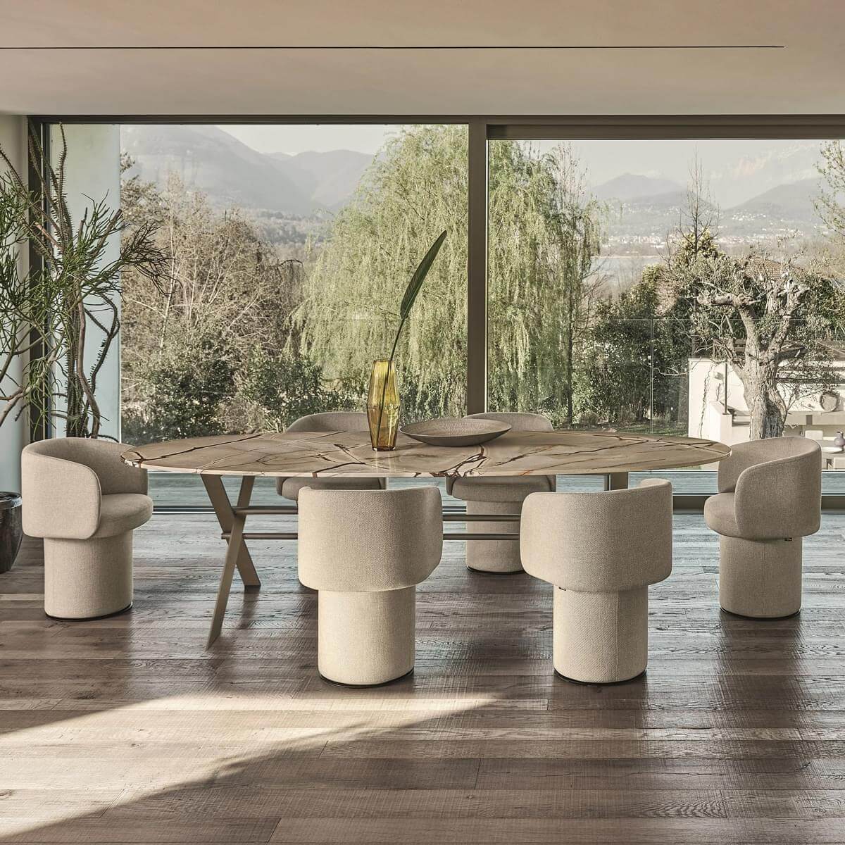 Marble dining table with grey veining, six beige upholstered armchairs, modern dining room with wood floors and landscape view.