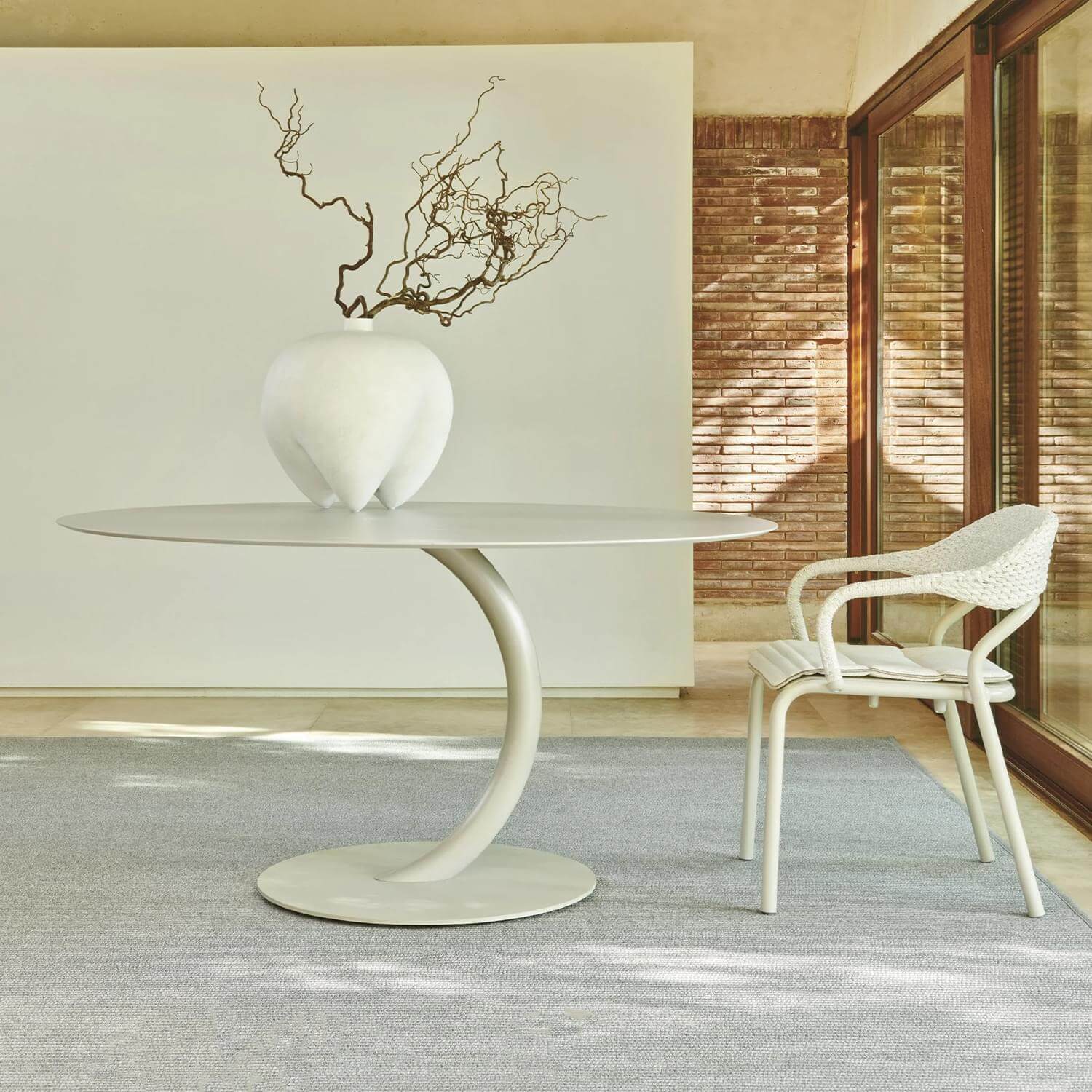 Round dining table with light gray concrete top and pedestal base, white woven chair, dried branches in vase.