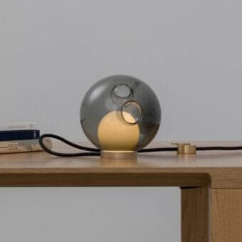 Grey translucent sphere on a light cylindrical base with a yellow illuminated inner shape and black cable on a wooden surface