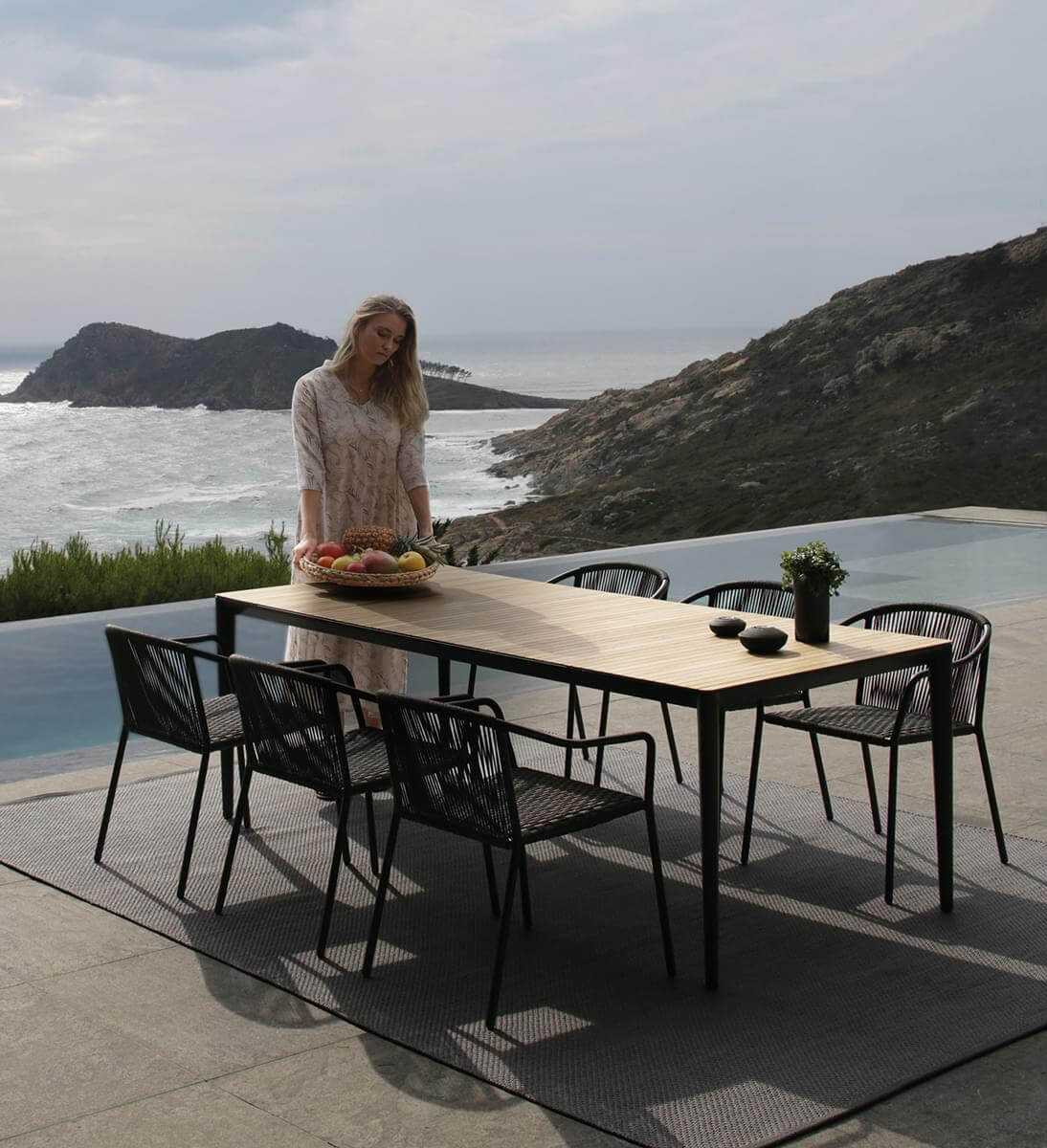 Outdoor dining table with light wood top and black metal frame, surrounded by six dark gray woven Samba armchairs on a gray tiled patio.